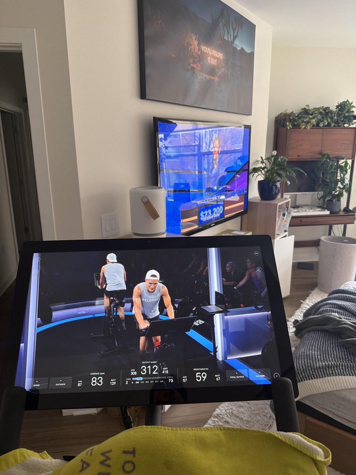 A Peloton screen mid-class showing live output, cadence, resistance and a power-zone bar, with Jeopardy playing on the television behind it.