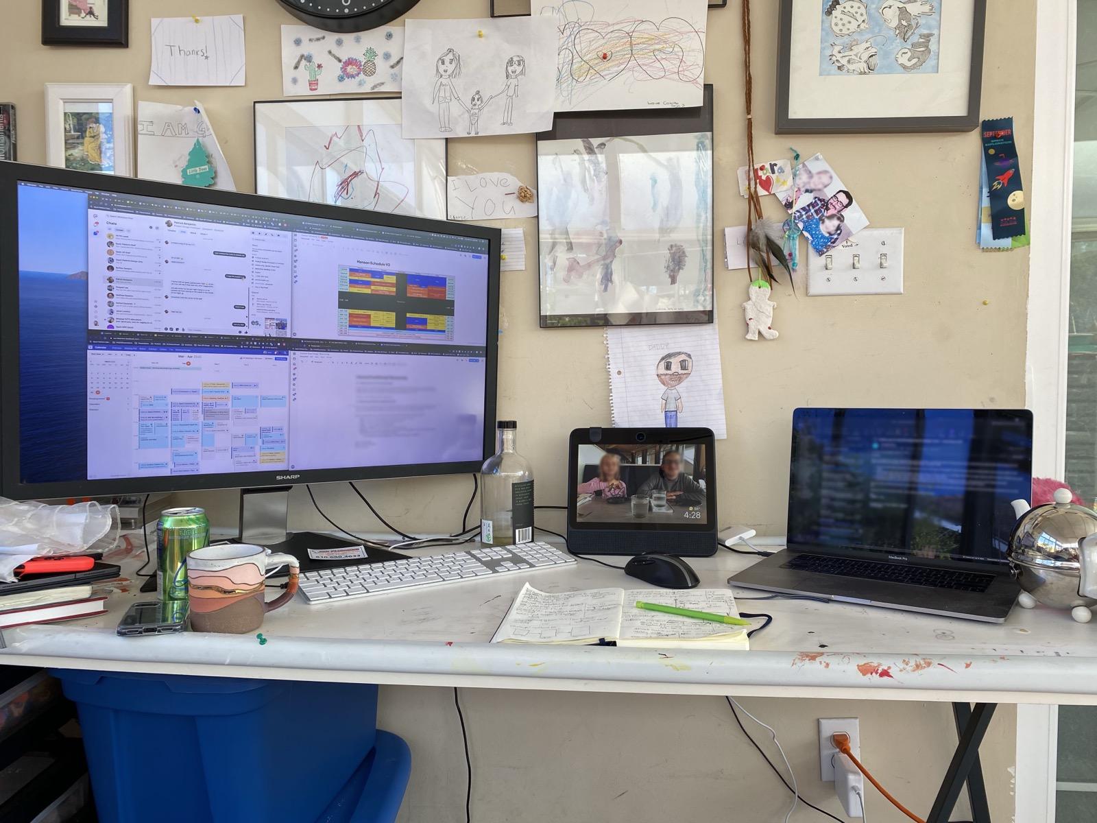 A home desk with a work monitor showing chat and a full calendar, a video call to family, and a wall covered in children’s drawings.